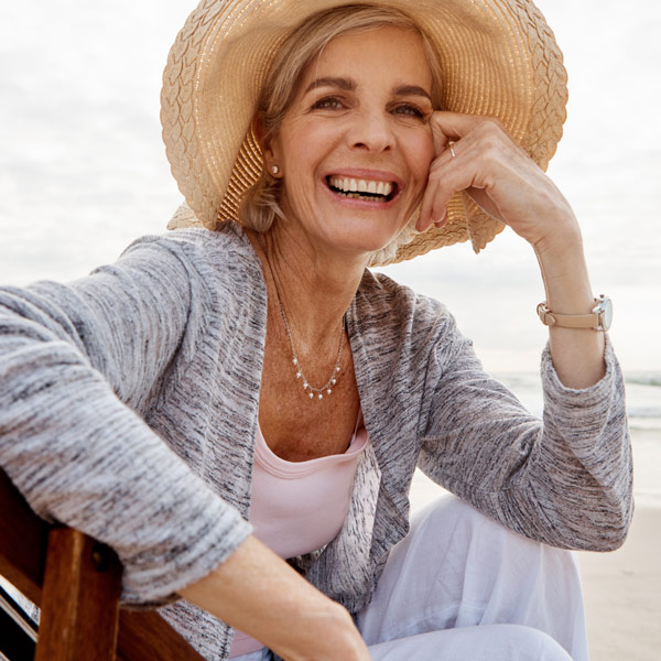 A senior woman wearing a hat, smiling while sitting on a beach in Santa Barbara, CA